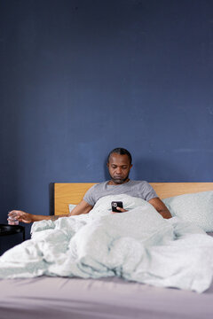 Man putting glass with water on table and using smartphone on bed