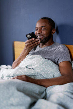 Pleased man recording audio message or talking on smartphone in bed