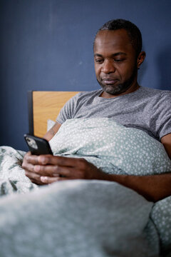 Black man using and watching smartphone lying under blanket on bed
