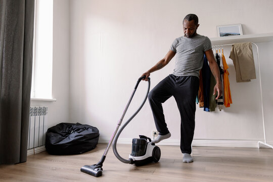 Black man pushing button of vacuum cleaner with leg in living room