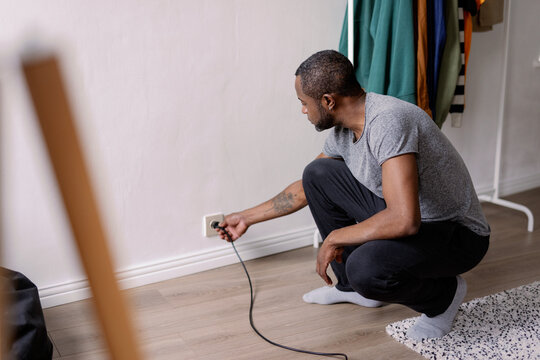 Adult black man plug on wire in outlet in room at home at daytime