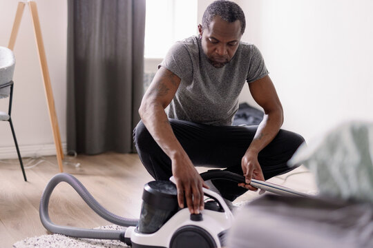 Adult black man pushing button of vacuum cleaner over carpet at home