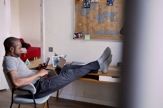 Side of focused adult black man using smartphone and laptop at home