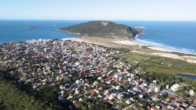 High-altitude aerial panorama of Santinho neighborhood in Florianopolis, Brazil, showcasing coastal houses, white sand dunes and the Morro dos Ingleses mountain under a clear blue sky.
