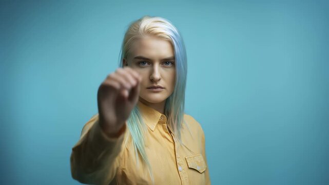 Serious young woman with dyed blue hair showing a stop gesture with her hand against a blue studio background