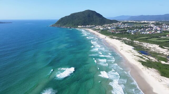 Low-to-high aerial tilt-up shot of Santinho Beach in Florianopolis, Brazil, revealing turquoise ocean waves, white sand dunes and the lush Morro das Aranhas mountain on a sunny day.
