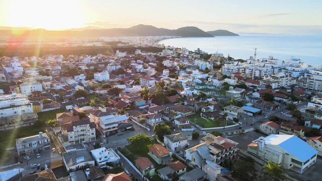 Cinematic sunset aerial panorama of Santinho and Ingleses neighborhoods in Florianopolis, Brazil, featuring coastal houses, residential buildings and the ocean under a golden sky.
