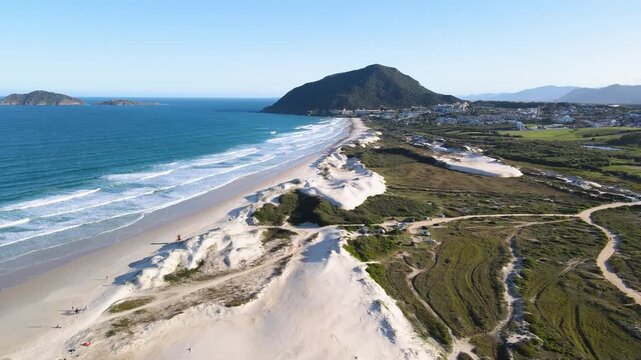 Fast low-altitude aerial hyperlapse over Santinho Beach dunes in Florianopolis, Brazil, flying towards Morro das Aranhas mountain with ocean waves and white sand.
