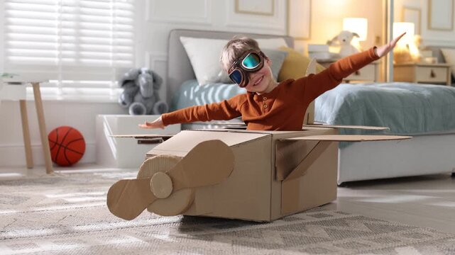 Happy little boy in cardboard airplane and goggles playing pilot on floor at home. Camera moving forward and backwards