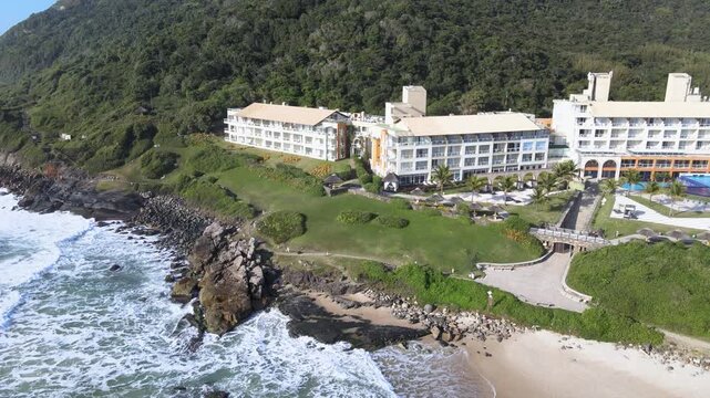 Cinematic aerial view of the luxury Costao do Santinho Resort in Florianopolis, Brazil, showing hotel buildings by the ocean waves and green mountains on a sunny day.
