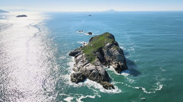 Cinematic high angle aerial shot of Ilha do Badejo in Florianopolis, Brazil, showcasing rocky cliffs, sun glint on turquoise water and fishing boats in the Atlantic Ocean.
