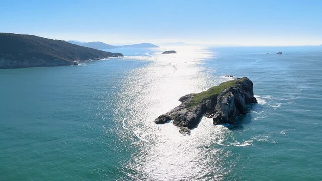 High-angle aerial shot of Ilha do Badejo in Florianopolis, Brazil, featuring rocky cliffs in the Atlantic Ocean with fishing boats and sun glint on turquoise water.
