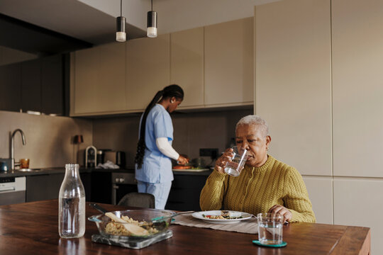 Senior woman drinking water while caregiver prepares meal in kitchen
