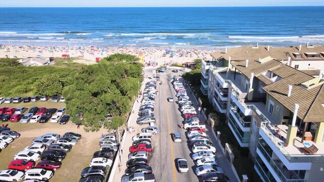High angle aerial view of Santinho Beach in Florianopolis, Brazil, featuring seaside apartments, crowded parking lots and tourists on the shore on a sunny summer day.
