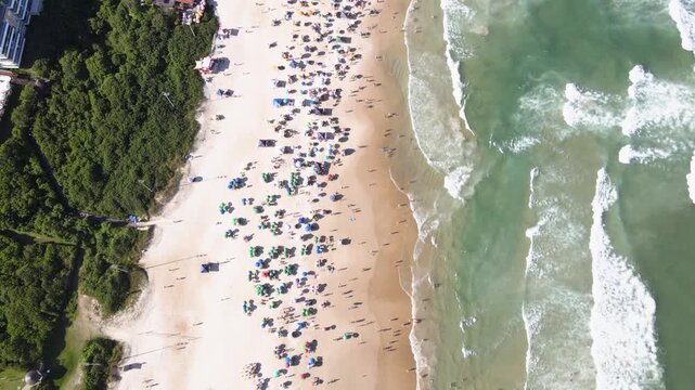Top down aerial view of a crowded Santinho Beach in Florianopolis, Brazil, featuring colorful umbrellas on white sand and turquoise ocean waves on a sunny summer day
