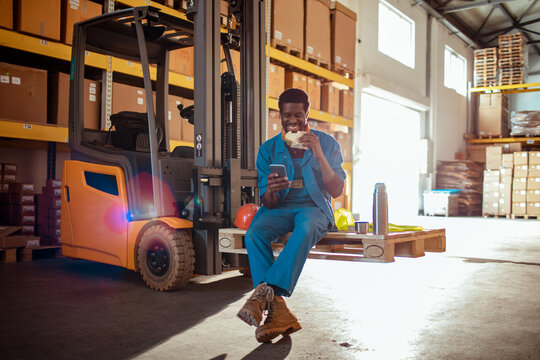 Warehouse worker eating lunch and using smartphone on forklift pallet