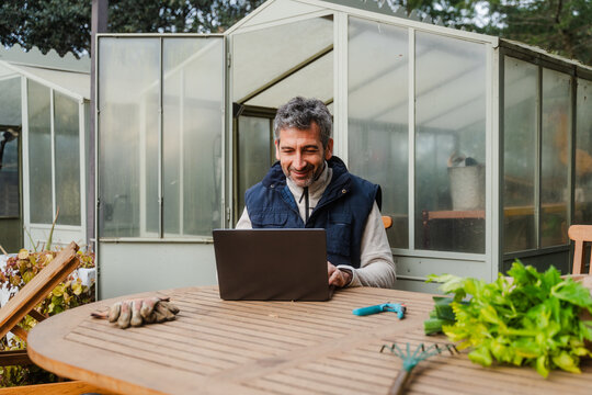 Male Owner Using Laptop While Sitting At Table In Greenhouse
