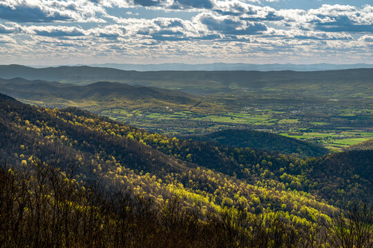 Sunlit spring foliage and rolling ridges above the Shenandoah Valley, with layered hills, farms, and dramatic broken clouds along the Blue Ridge Parkway.