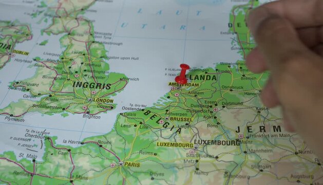 A man's hand is sticking a red pushpin into Amsterdam is the capital and largest city of the Netherlands, on the geographical map of Europe