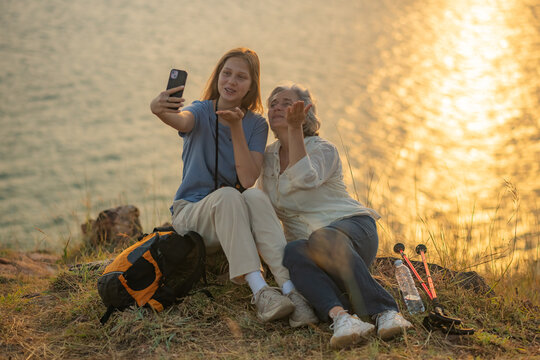 Hiker on grassy hill overlooking sea sunset taking photo with her smartphone backpack and trekking beside representing travel adventure and peaceful outdoor lifestyle.