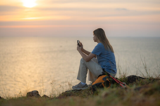Hiker on grassy hill overlooking sea sunset taking photo with her smartphone backpack and trekking beside representing travel adventure and peaceful outdoor lifestyle.