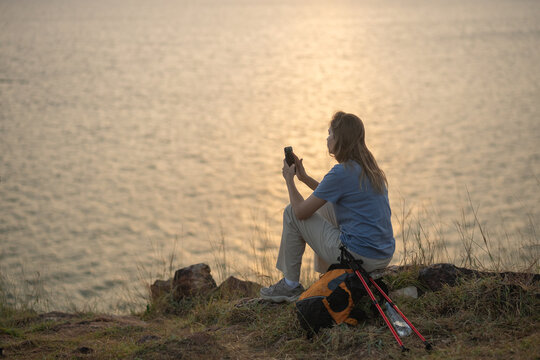 Hiker on grassy hill overlooking sea sunset taking photo with her smartphone backpack and trekking beside representing travel adventure and peaceful outdoor lifestyle.