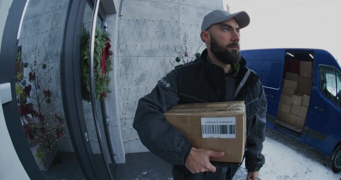 Bearded Courier Hands Over Parcel and Manages Electronic Signature Process on Porch Decorated With Christmas Tree. Concept of Busy Holiday Shipping Season, and Modern Contactless Ecommerce Logistics.