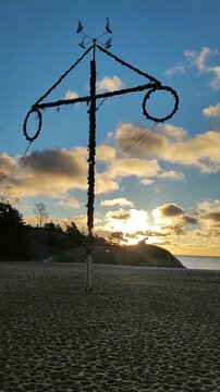 Traditional Midsummer Maypole on a winter sandy beach waiting for spring. A tall maypole decorated with greenery stands against a against the backdrop of the blue sky and the setting sun. 