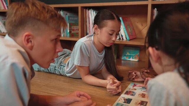 Smart girl looking at picture book while boy talking with diverse friend at library. Cute high school student lying down in circle near bookshelf while group of children sharing book. Edification