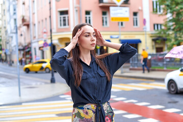 Fototapeta premium A woman in a black shirt and trousers decorated with a floral pattern stands on a city street. Her gaze is fixed on the camera with a serious expression on her face. Tall buildings line the sidewalk.