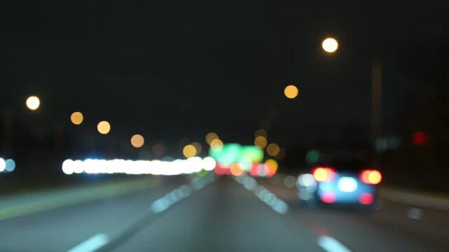 Blurred POV View of multiple cars driving on a highway at night with glowing headlights and taillights. Scene shows urban transportation, night traffic. 