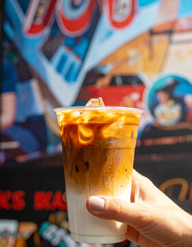Fresh iced caffe latte featuring beautiful espresso layers and milk. Hand-held in a clear cup against a vibrant mural background. Perfect for a refreshing caffeine kick.