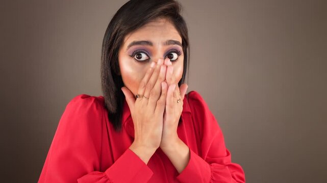 Close up of shocked surprised attractive young Asian woman lady wear red shirt looking at camera covering mouth with hands feels astonishment advertising, isolated over grey studio background. 