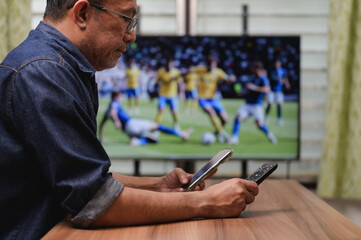 World cup 2026, Asian man holding smartphone remote control while watching soccer match on television at home, checking live scores betting online during intense football game competition. © Mdisk