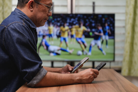 World cup 2026, Asian man holding smartphone remote control while watching soccer match on television at home, checking live scores betting online during intense football game competition.