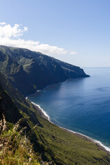Ponta do Pargo Atlantic cliffs Madeira Portugal vertical coastline view, steep green volcanic slopes descend toward calm blue ocean with narrow surf edge and vast clear sky