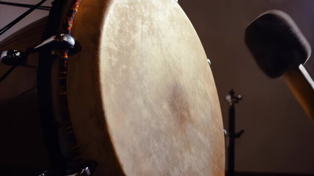 Close-up of a hand striking a drum with a mallet, capturing the motion and impact on the drum surface in a softly lit indoor setting