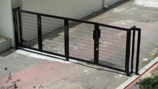 A modern black metal folding gate with a mesh design secured on a concrete driveway. An architectural detail focusing on urban residential security, property boundaries, and gate design.
