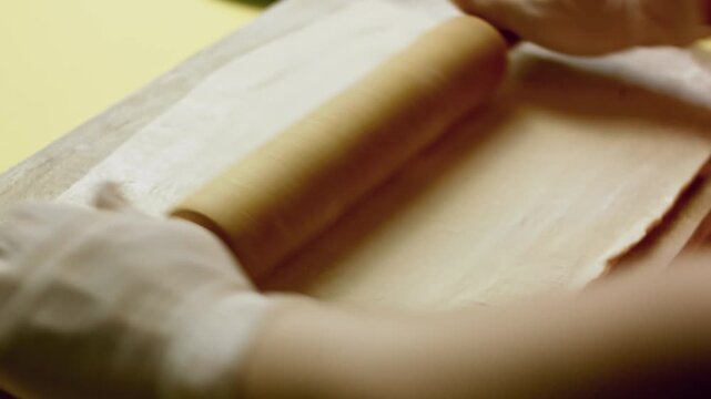 Wooden rolling pin is being used to flatten a sheet of puffy pastry