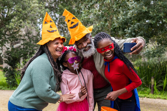 African American family posing in park while senior man and girls taking phone selfie, pumpkin hats