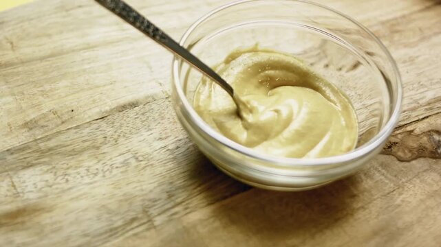 Glass bowl with a spoon containing Dijon mustard on a wooden cutting board