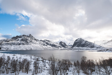 Obraz premium Sunlight breaks through heavy clouds over the snowy fjords near Sund, Lofoten, Norway, illuminating a lone red cabin in a dramatic Arctic winter landscape