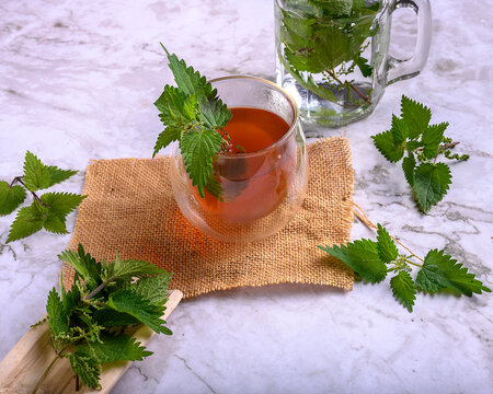 Nettle herbal tea in a double wall glass on burlap and marble surface, fresh stinging nettle leaves around a jar with water, concept of healthy detox beverage and natural medicine