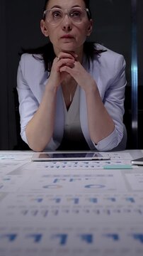 Businesswoman intently reviewing financial charts and reports scattered across her desk while using a digital tablet, working overtime in a corporate environment