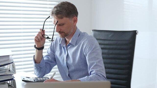 Male professional accountant wearing blue shirt and glasses is calculating invoices and managing bills while reviewing financial reports in an office setting. Audit and taxes theme in business