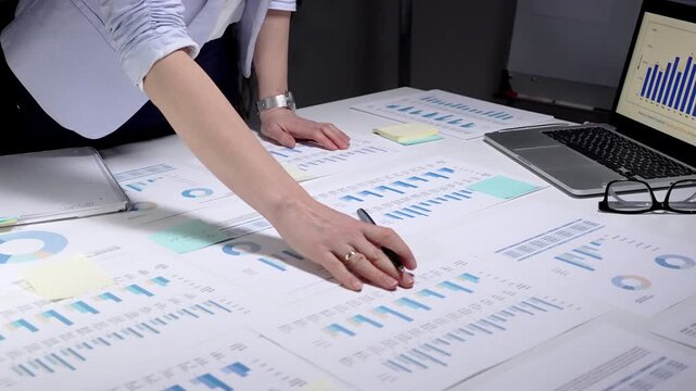 Professional businesswoman working at a desk surrounded by financial reports, charts, and graphs, focusing on data analysis to develop strategic plans and improve performance