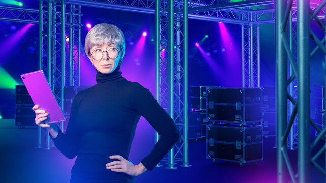 Event production, Stage management. Female technical coordinator plans lighting and equipment setup using tablet at concert venue with truss structures and stage cases.