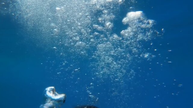 Cinematic footage of a scuba diver wearing a mask and exhaling air bubbles while diving deep in the clear blue ocean water in slow-motion.