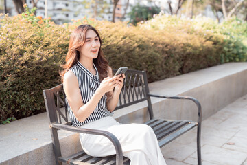 Obraz premium Young Asian woman sitting on a metal bench in a park, smiling and actively using her mobile phone for communication, social media, or online tasks during a sunny day