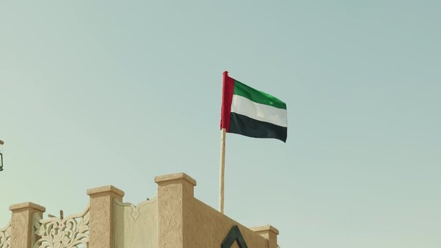 National flag of the United Arab Emirates, with red, green, white, and black colours, waving in the wind against a clear blue sky.